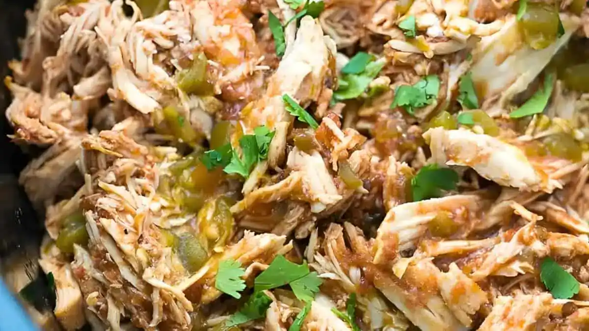 A close-up shot of shredded chicken mixed with chopped green chilies, herbs, and spices. The seasoned meat displays a blend of vibrant colors and textures, topped with fresh cilantro leaves.