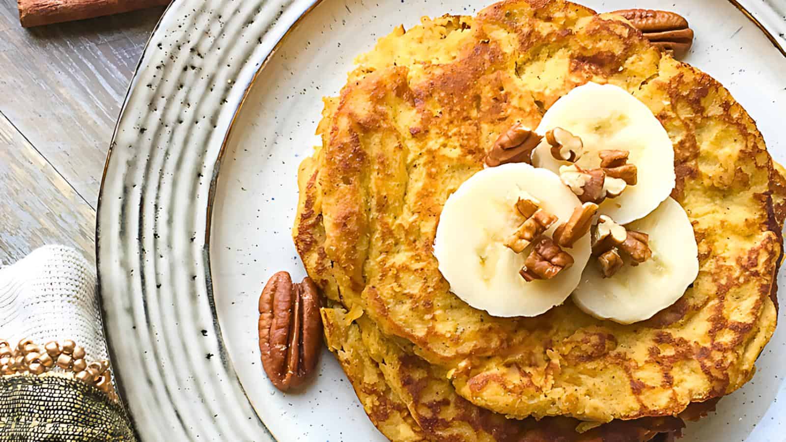A plate of golden brown pancakes topped with banana slices and chopped pecans. A few whole pecans are placed next to the pancakes on a white and beige plate. The dish is set on a light wooden surface with a decorative white and beige napkin nearby.