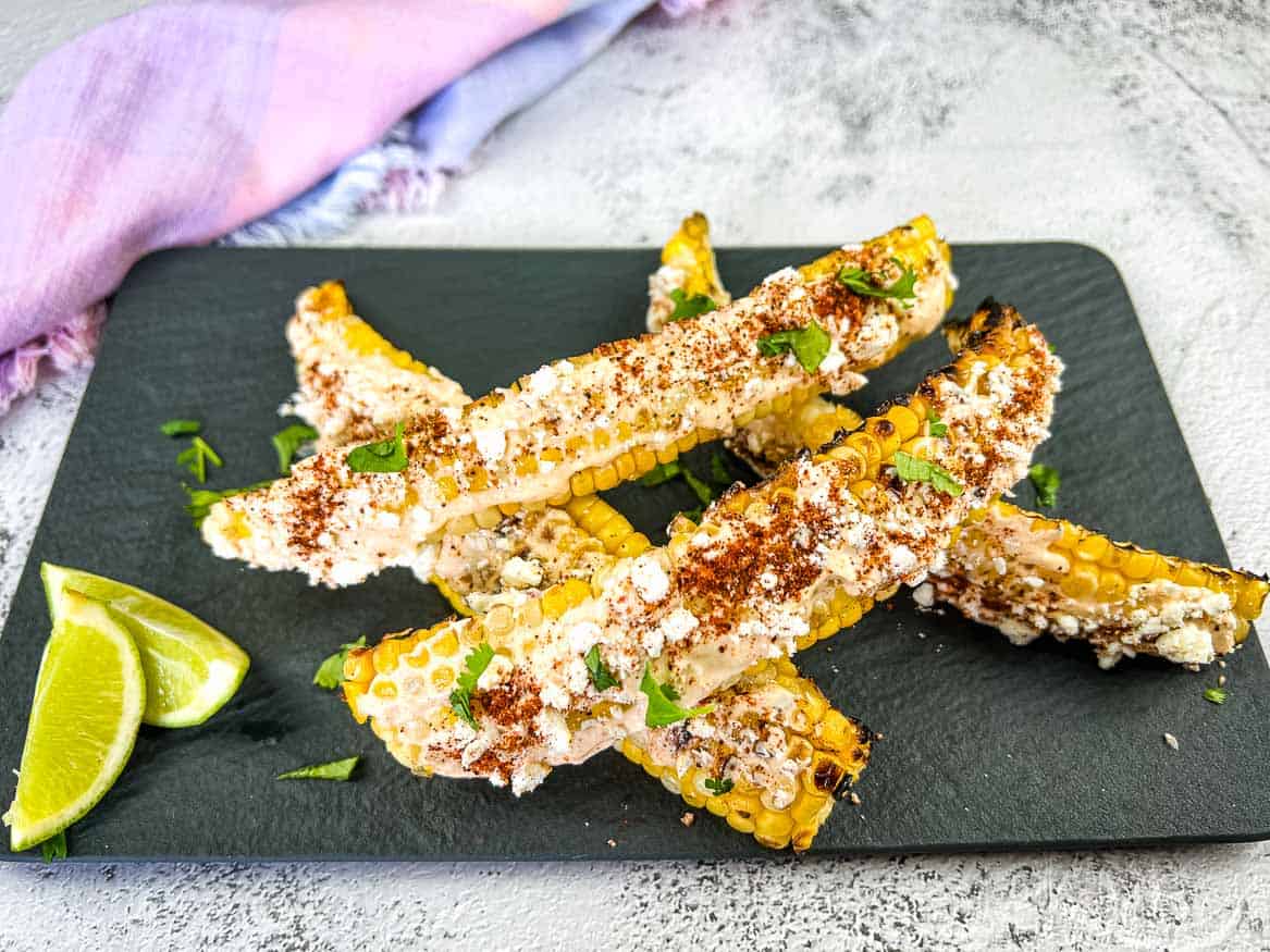 A gray slate plate holds several pieces of Mexican street corn, or elote, coated with mayonnaise, cheese, chili powder, and garnished with cilantro. Lime wedges are placed on the side. A pink and purple cloth is partially visible in the background.