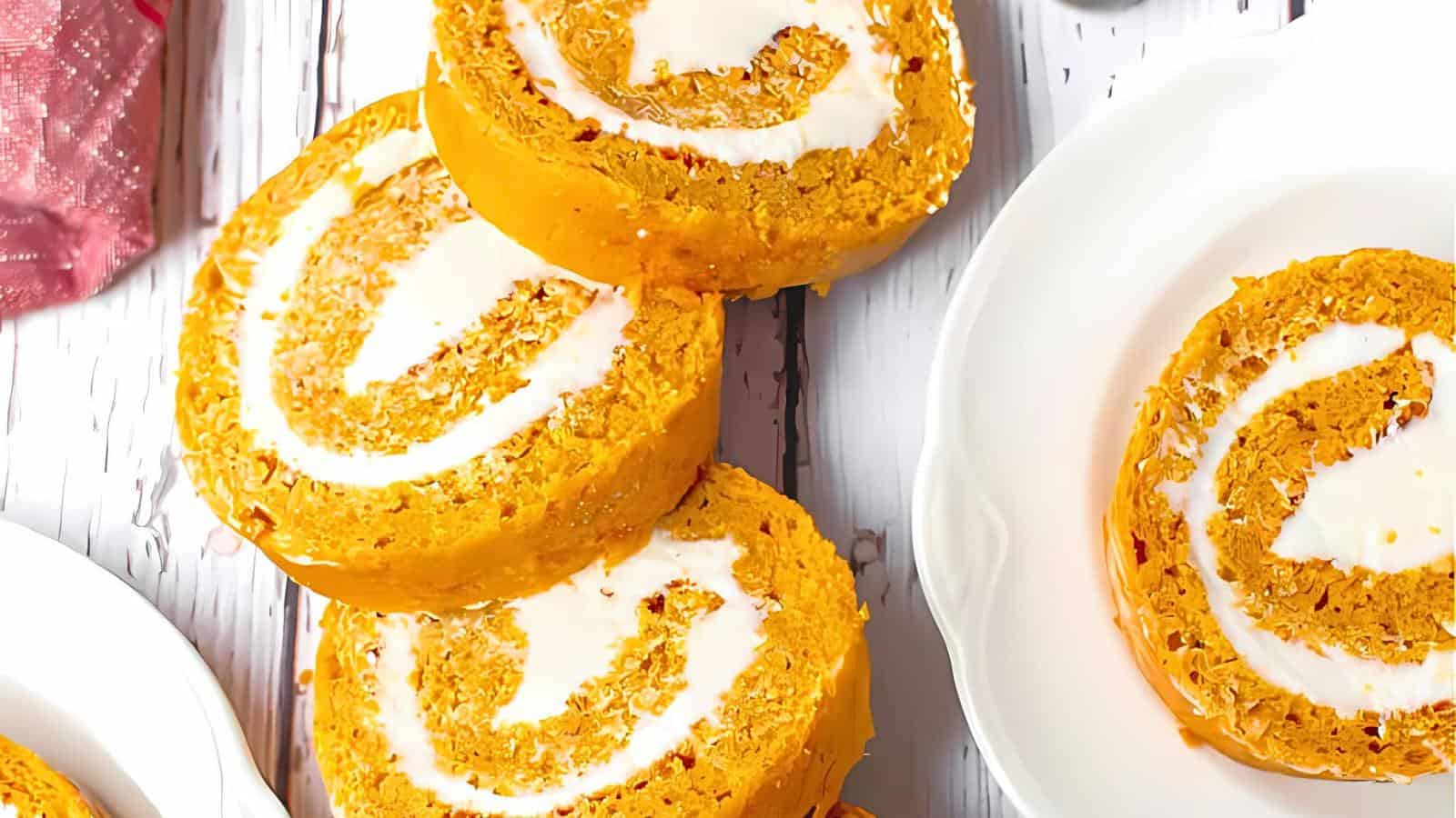Pumpkin roll slices with a creamy white filling are neatly arranged on a white rustic wooden surface, with one slice placed on a white plate. A pink sparkly cloth is partially visible on the left side of the image, adding a touch of color.