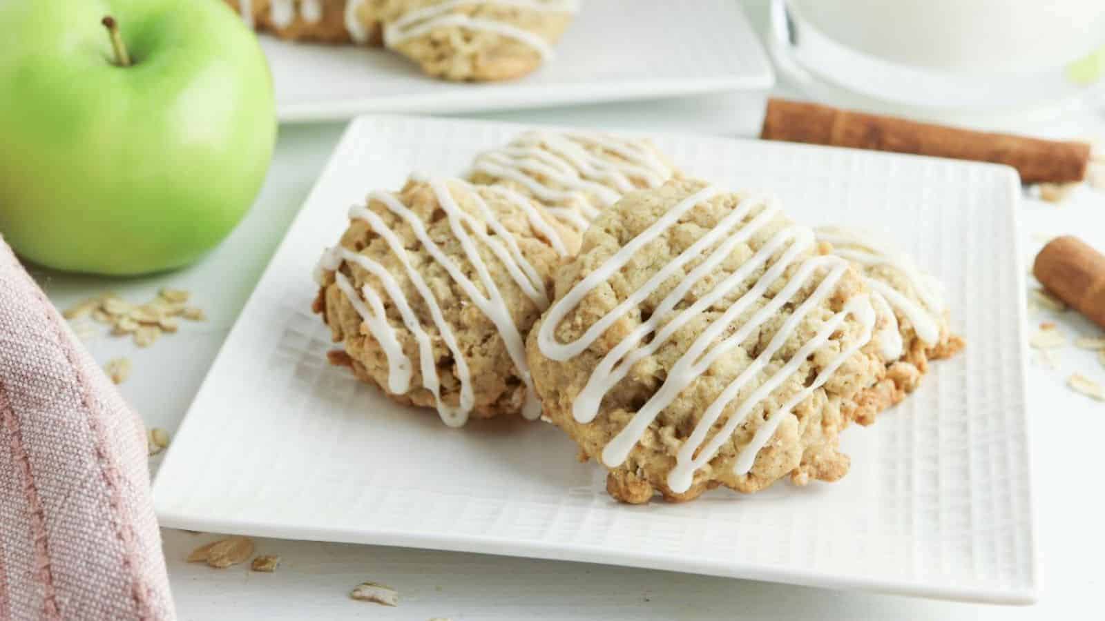 Three apple oatmeal cookies drizzled with white icing are placed on a white square plate. In the background, a green apple, cinnamon sticks, some scattered oats, and a glass of milk are visible. The setting appears to be a clean, well-lit kitchen or dining area.