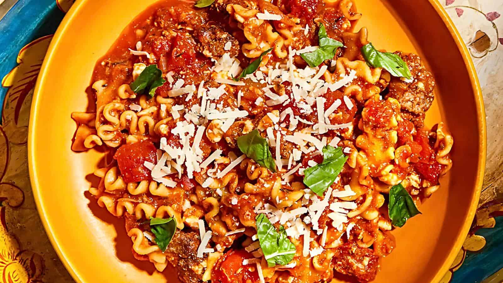 A vibrant dish featuring cooked pasta with a red tomato-based sauce, topped with grated cheese and fresh basil leaves. The pasta is served in a yellow bowl, showcasing a hearty mixture of tomatoes and possibly meat pieces, garnished for added color and flavor.