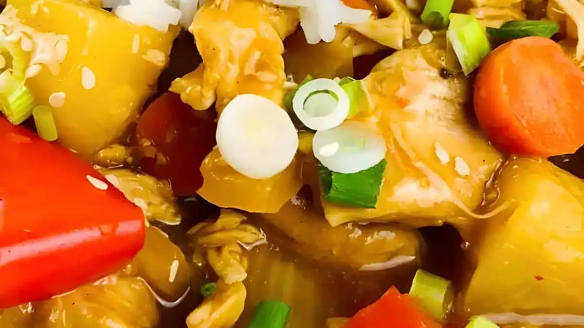 Close-up of a colorful stir-fry dish, featuring chunks of chicken, pineapple, carrots, and red bell peppers in a glossy sauce. Garnished with sliced green onions and sesame seeds. The vibrant colors and textures showcase a fresh and appetizing meal.