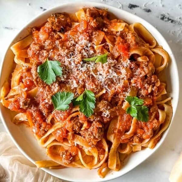 A white bowl of tagliatelle with meat tomato sauce, grated cheese, and parsley on marble next to a block of cheese and a fork.