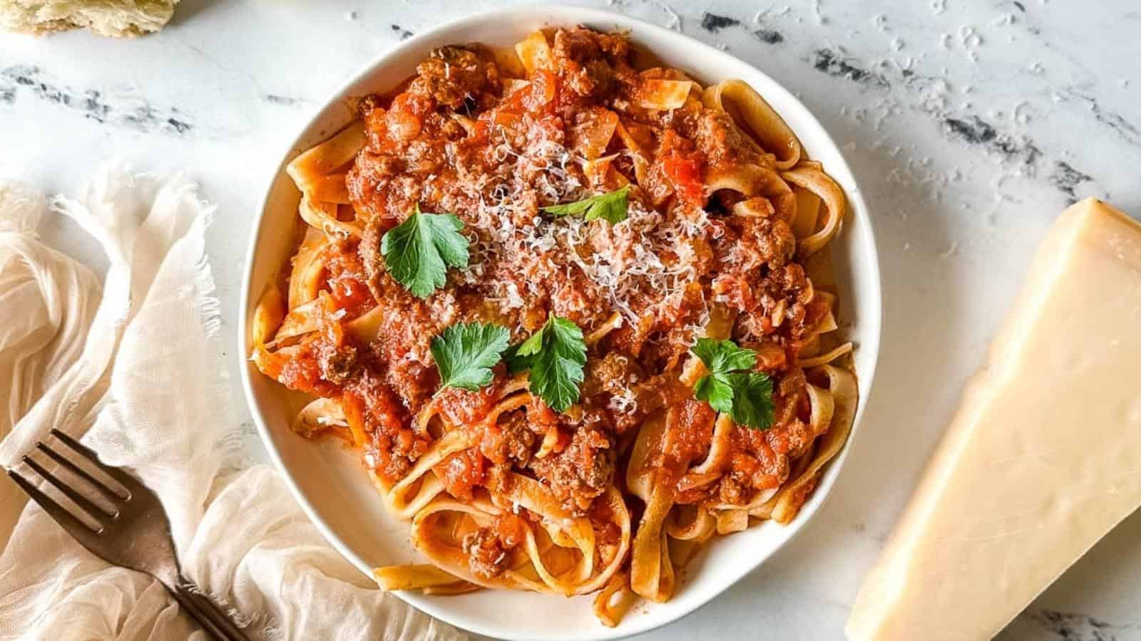 A white bowl filled with pasta topped with a rich red tomato meat sauce, garnished with fresh parsley and grated cheese. Beside the bowl, there is a fork resting on a beige cloth napkin and a block of Parmesan cheese on a marble countertop.