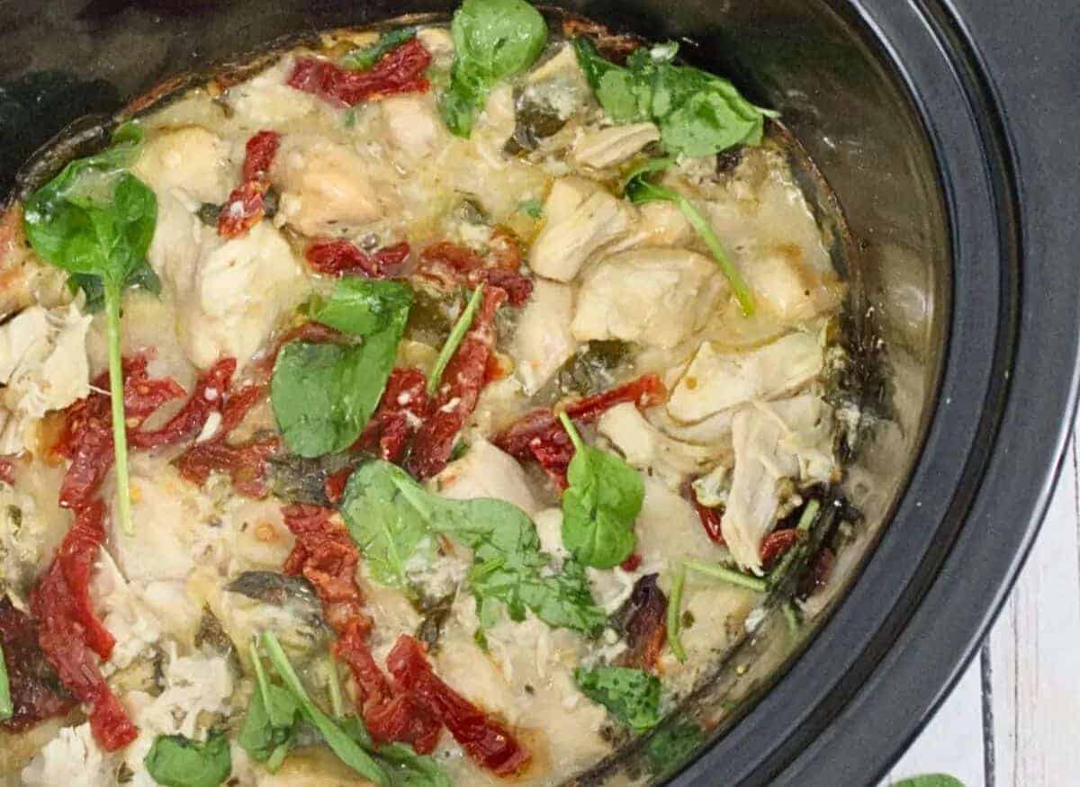 A close-up of a crockpot dish featuring creamy chicken with wilted spinach and sun-dried tomatoes. The chicken is shredded and mingled with the spinach, creating a colorful and hearty meal with a creamy consistency.