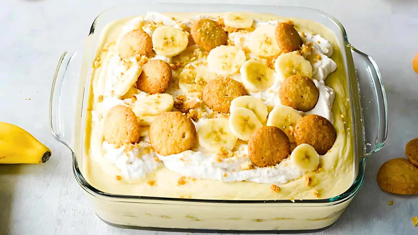 A square glass dish holds a banana pudding with layers of vanilla wafers, sliced bananas, and whipped cream, topped with more banana slices and cookies. A whole banana and some vanilla wafers are visible beside the dish.