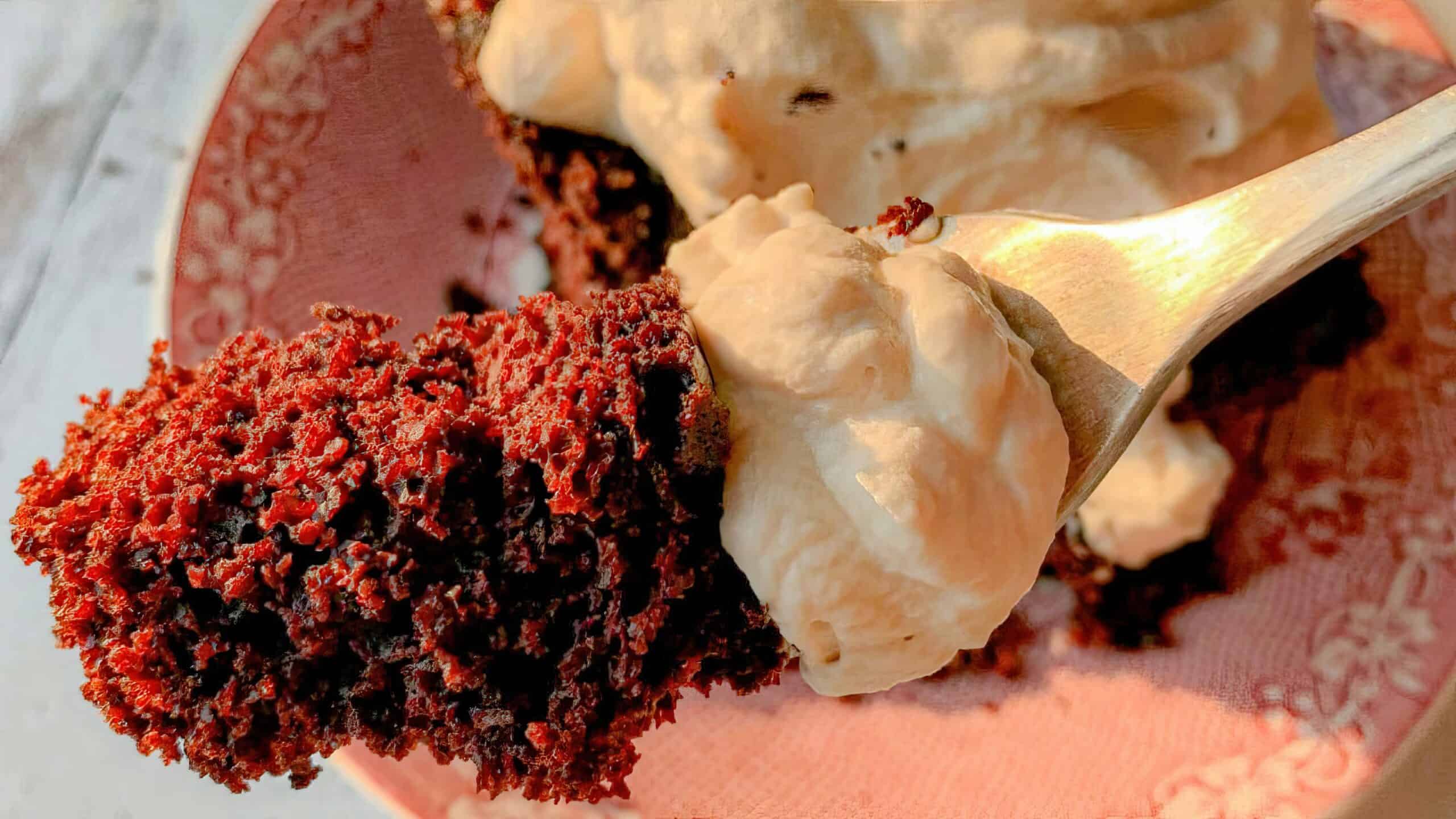 A close-up of a fork holding a piece of red velvet cake with a generous dollop of cream cheese frosting. The cake has a rich, moist texture and deep red color, contrasting with the creamy, smooth frosting. The plate below is pink with a delicate floral pattern.