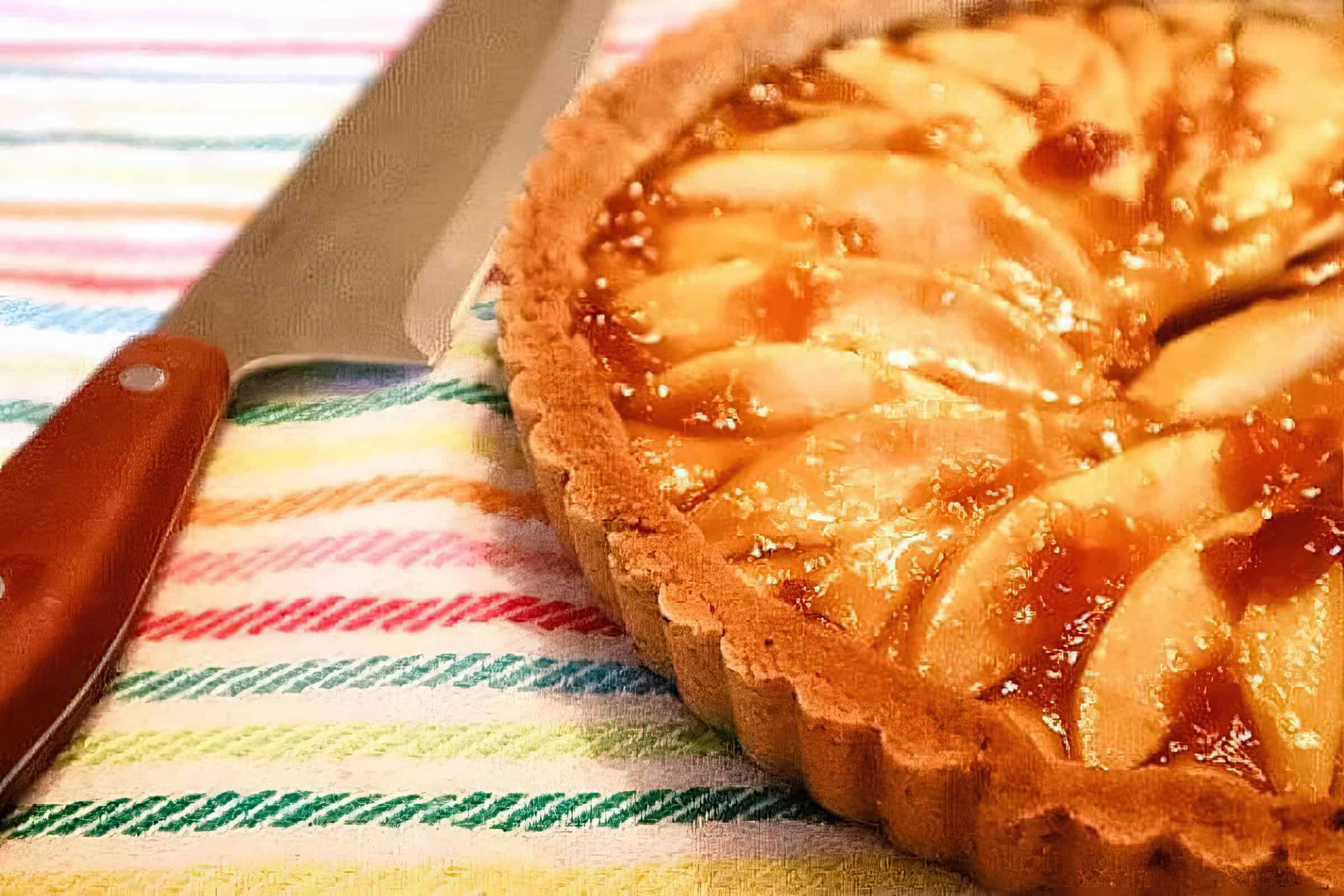 A close-up of a freshly baked apple tart with a golden crust and caramelized apple slices arranged in a circular pattern on top. The tart is placed on a colorful, striped cloth with a knife beside it.