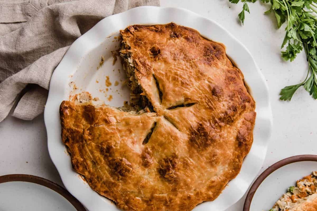 A golden-brown pie with a slice missing is displayed in a white ceramic pie dish. The pie's flaky crust is marked with four slits in the center. Fresh parsley and a beige cloth napkin are visible in the background.