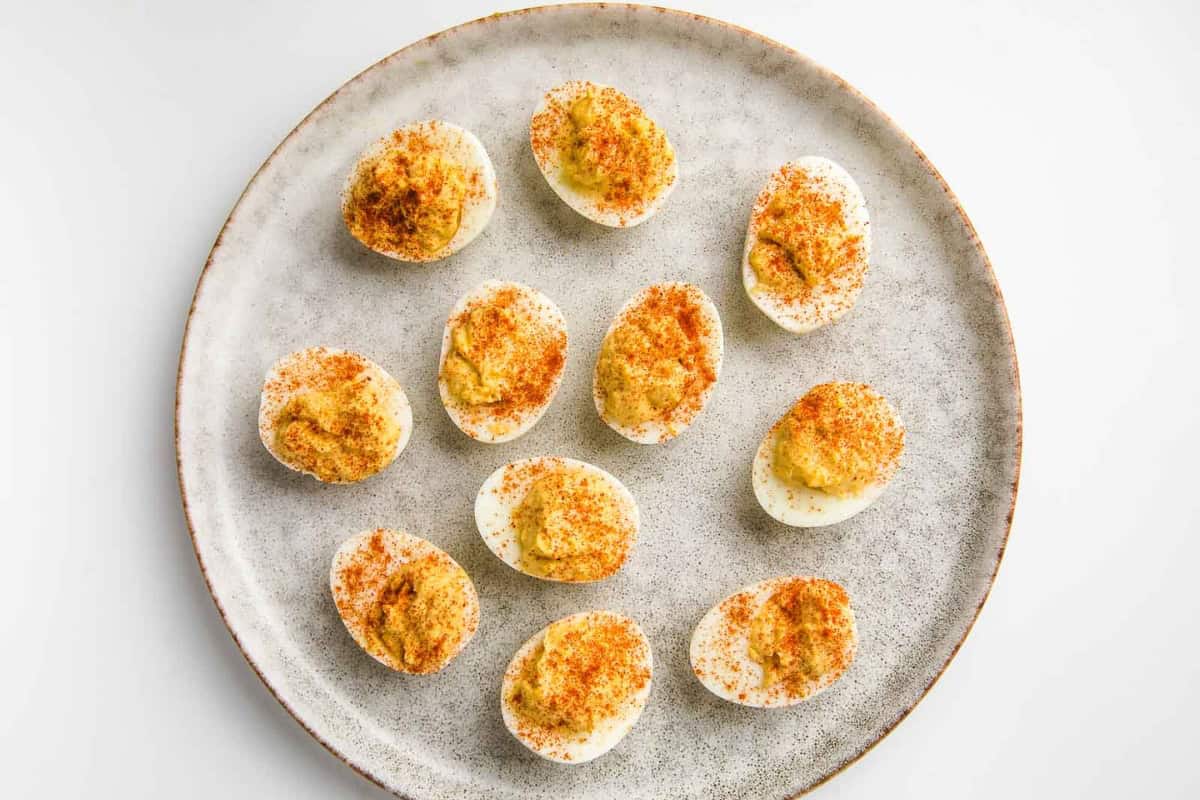 A round plate features a neatly arranged selection of deviled eggs. Each halved egg is filled with a creamy yolk mixture and topped with a sprinkle of paprika, creating a visually appealing appetizer. The background is a plain white surface.