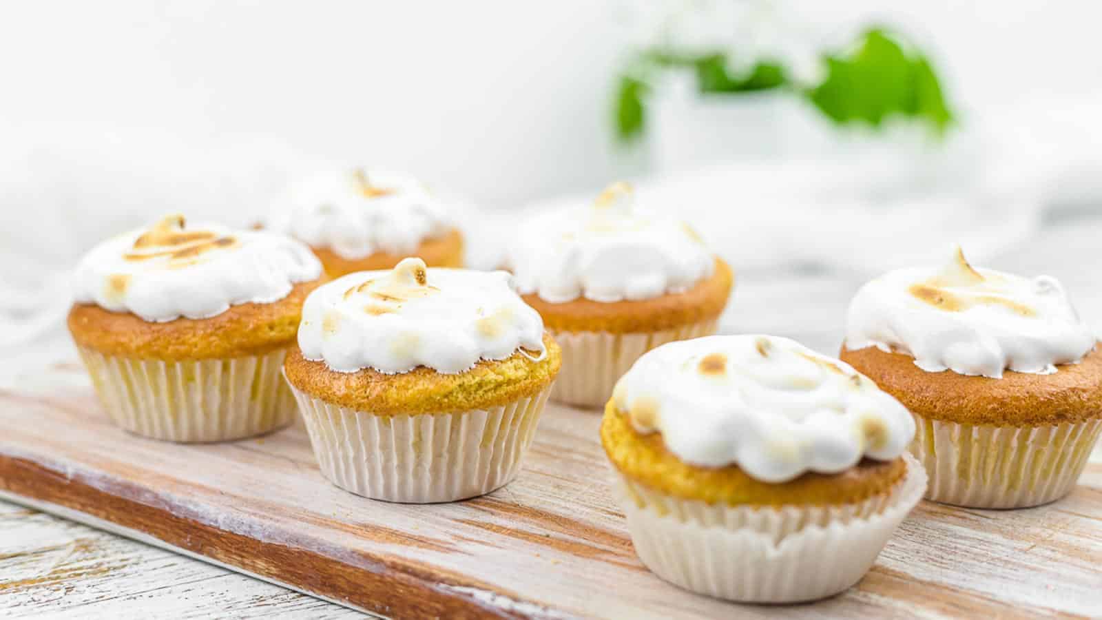 Seven cupcakes with golden-brown tops and white frosting, arranged on a wooden tray. They are in a bright setting with blurred greenery in the background, creating a fresh and inviting appearance.