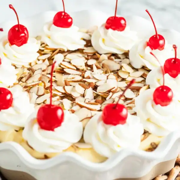 Whipped cream swirls, maraschino cherries, and sliced almonds top a pie in a white dish set on a wicker tray.