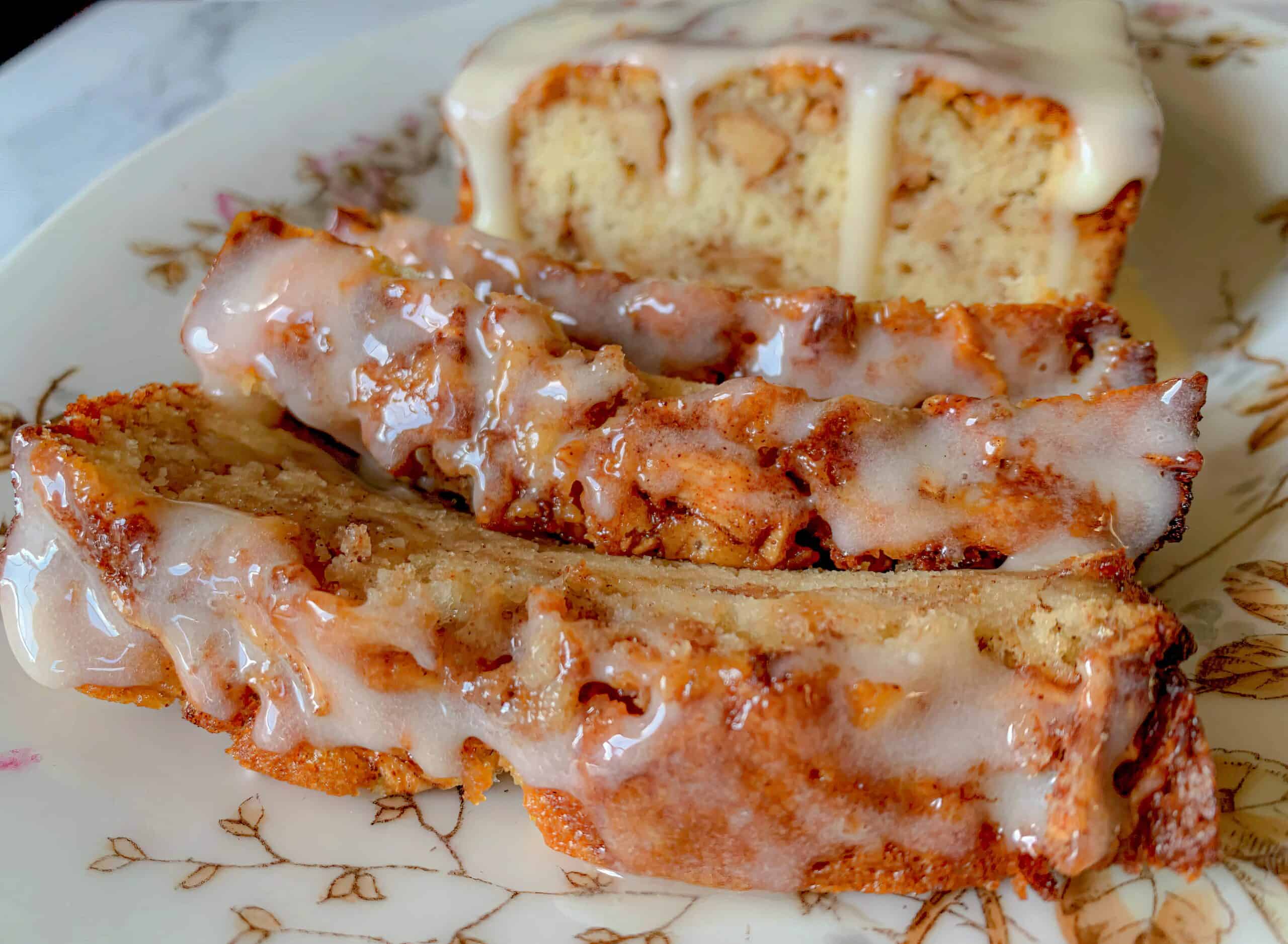 A close-up of a sliced cinnamon bread loaf with a glaze on top. The slices are arranged on a floral-patterned plate, showing the moist texture and the glistening glaze, making the dessert look delicious and appetizing.