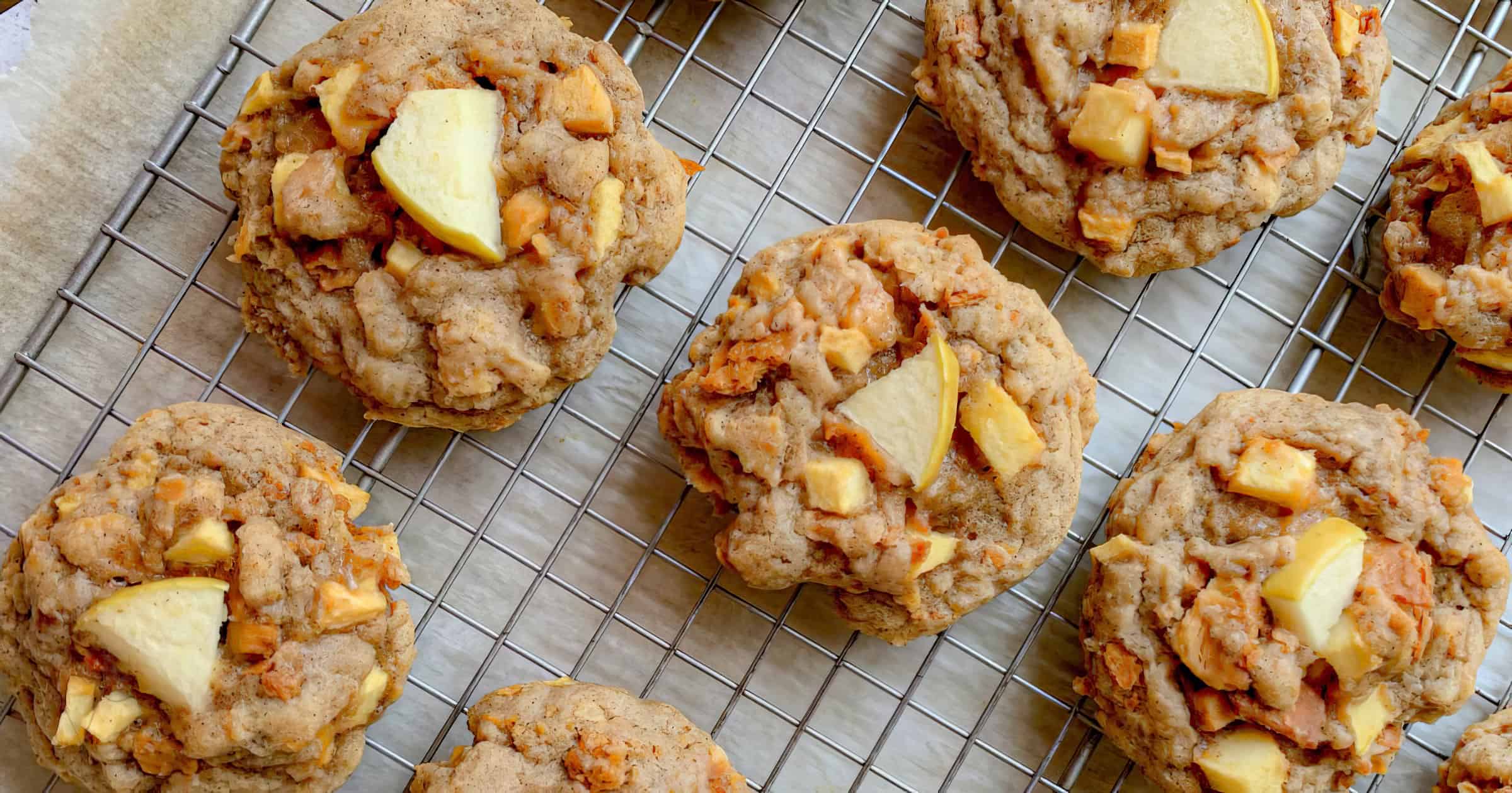 A cooling rack holds several freshly baked apple cinnamon cookies with chunks of apple and a golden-brown exterior. The rack is placed on a parchment paper-lined baking sheet.