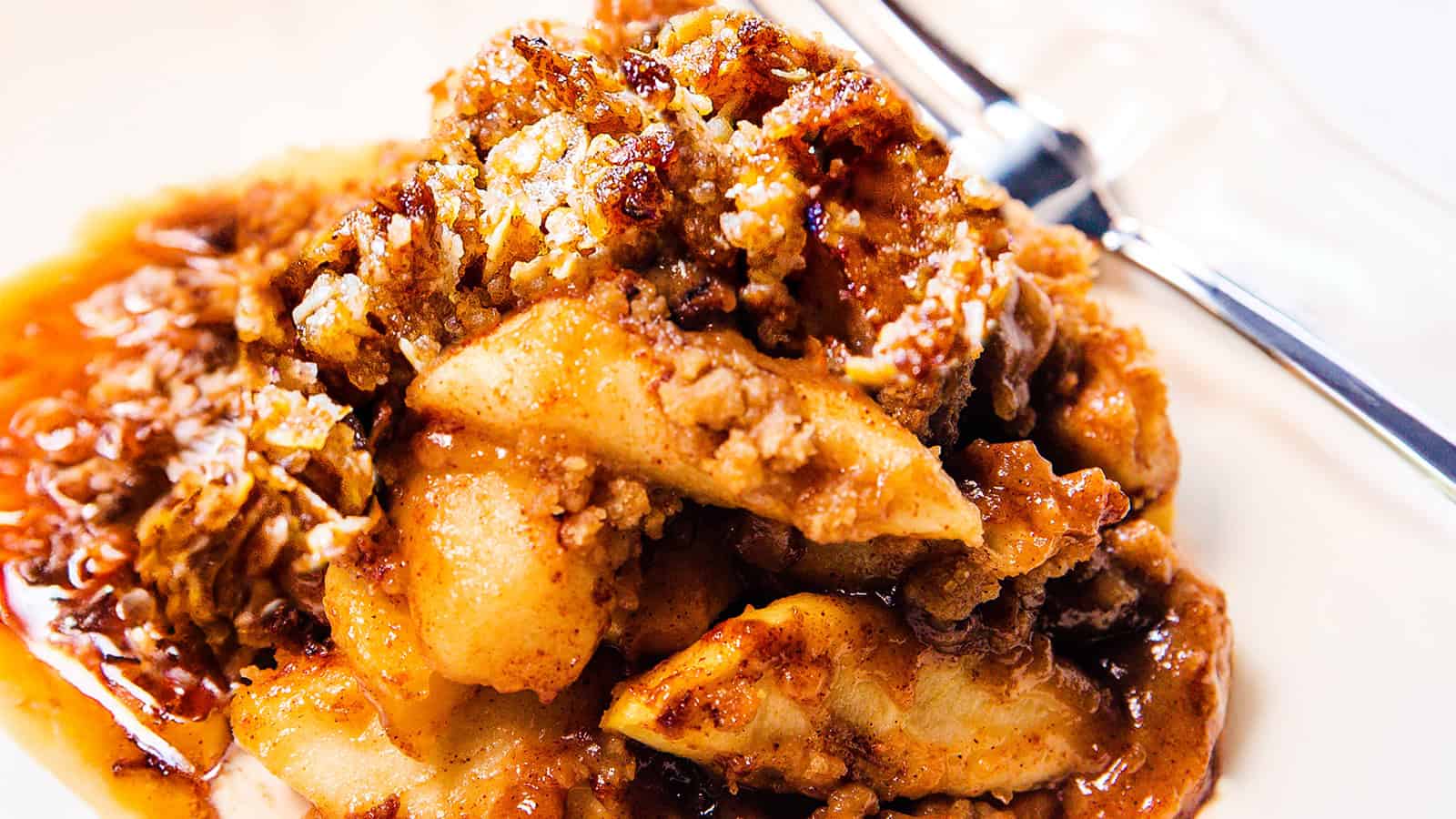 A close-up shot of a serving of freshly baked apple crisp, featuring caramelized apple slices topped with a golden-brown, crumbly streusel. A spoon, partially visible, rests on the side of the white plate. The dish looks warm and inviting.
