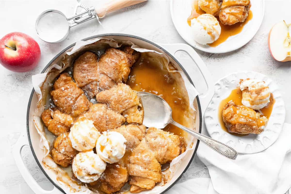 A white dish filled with freshly baked apple dumplings topped with scoops of vanilla ice cream and caramel sauce. A large serving spoon rests in the dish. Surrounding it are a red apple, a sifter, and a plate with apple dumplings and ice cream.