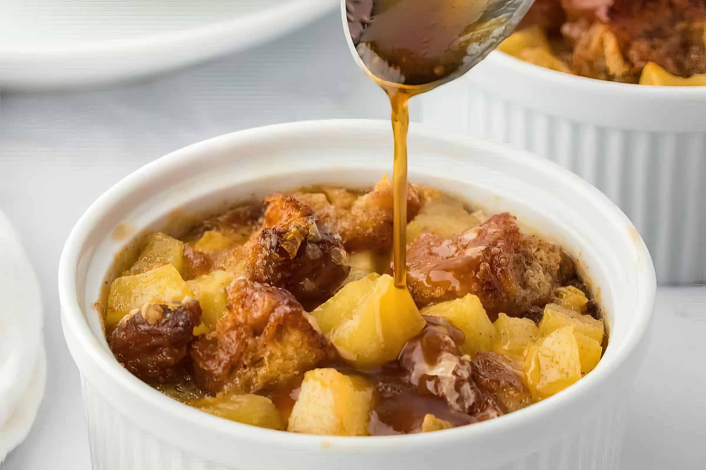 A spoon drizzles caramel sauce over a ramekin filled with bread pudding. The dish is topped with chunks of apple and raisins. The ramekin sits on a white surface, and another similar dish is partially visible in the background.