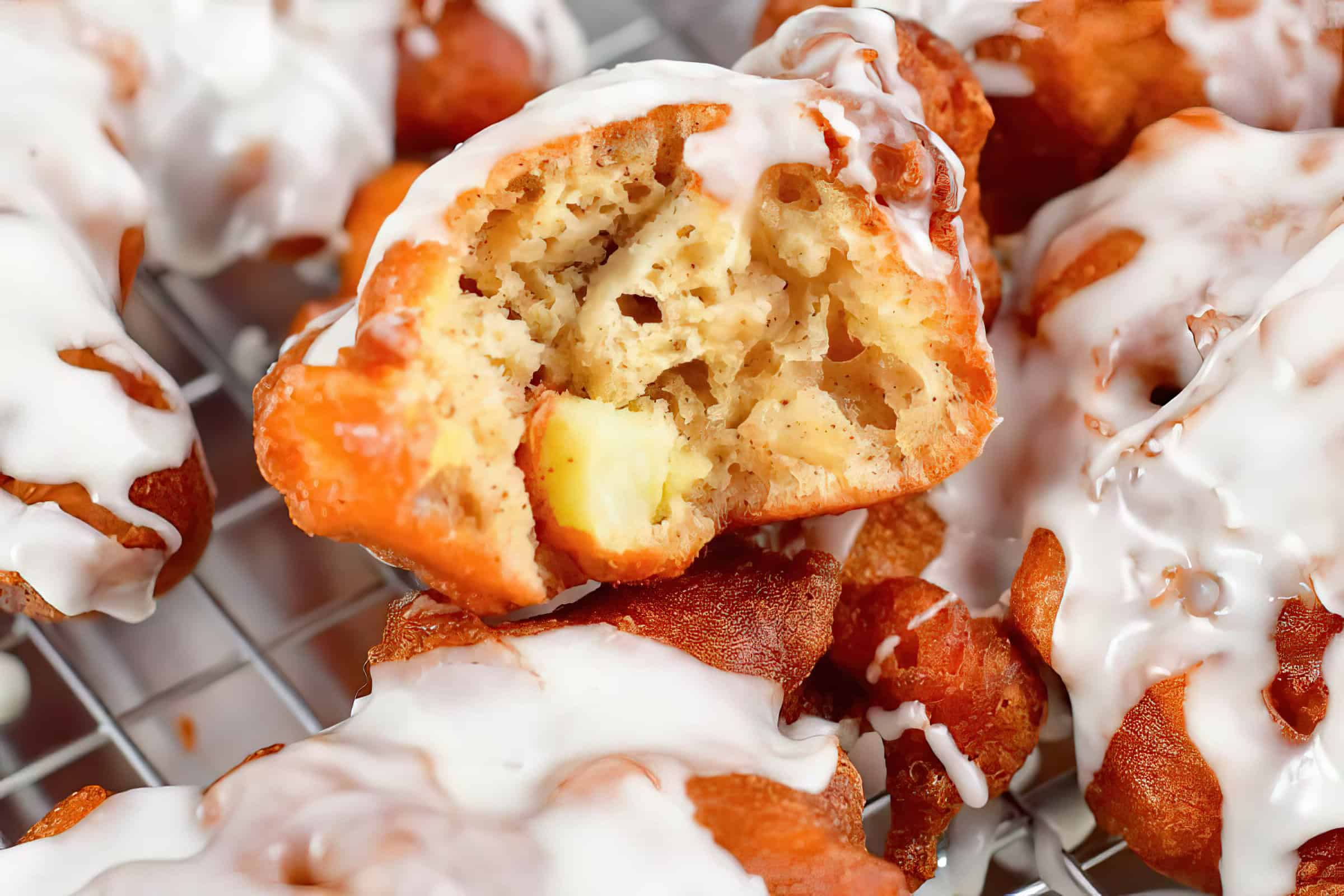 Close-up of a delicious apple fritter with a bite taken out, showcasing its moist interior. The fritter is generously drizzled with a sweet glaze and sits on a cooling rack alongside other similarly glazed fritters.