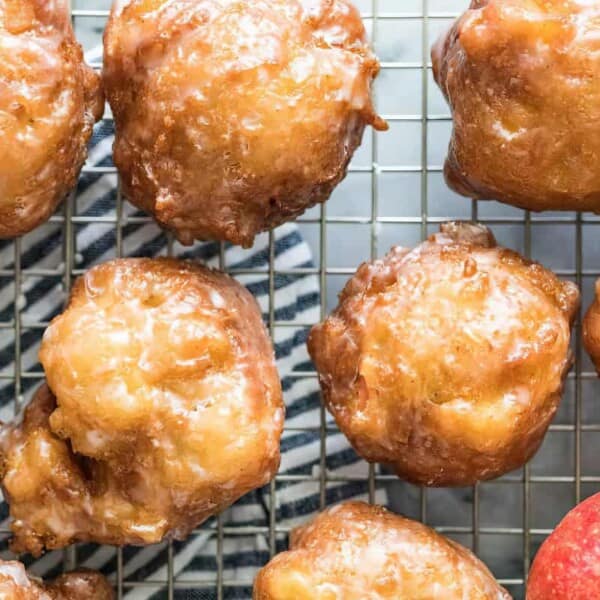 A close-up of several glazed apple fritters cooling on a wire rack. The fritters are golden-brown and shiny with glaze. A red apple is visible in the bottom right corner, and a patterned cloth is partially visible under the wire rack.