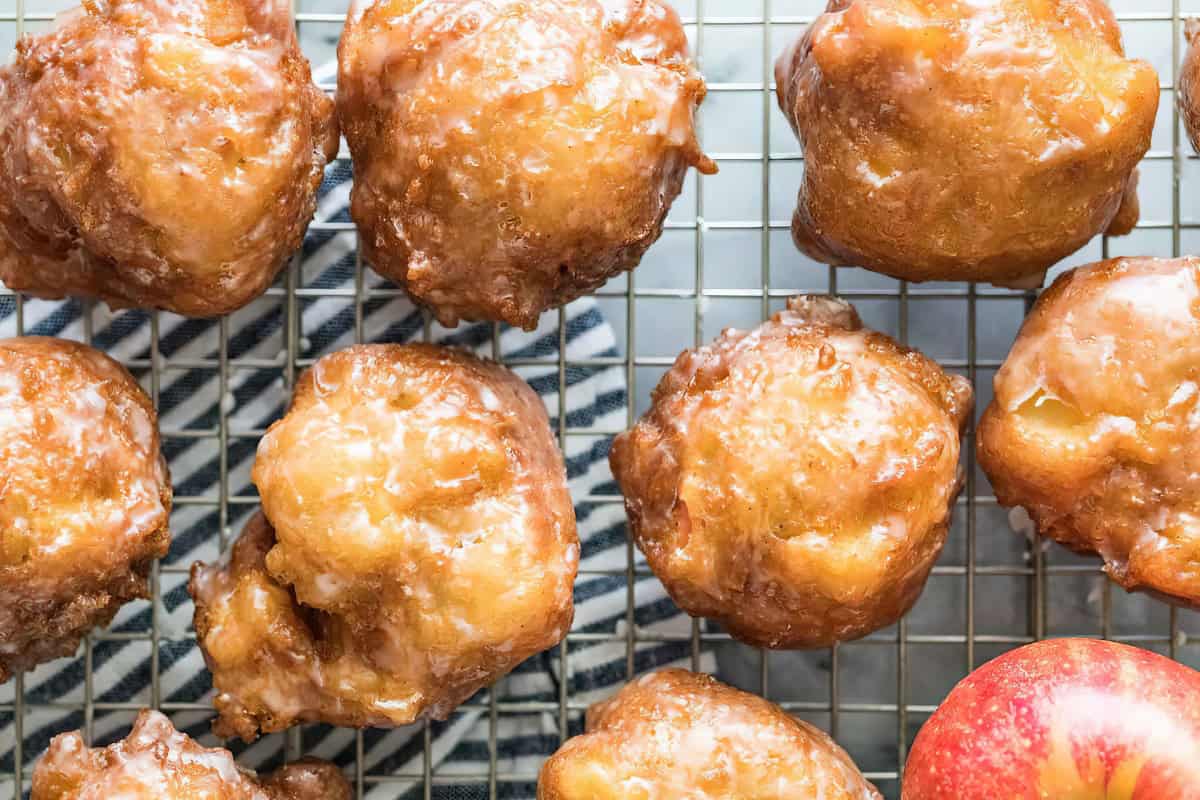 A close-up of several glazed apple fritters cooling on a wire rack. The fritters are golden-brown and shiny with glaze. A red apple is visible in the bottom right corner, and a patterned cloth is partially visible under the wire rack.