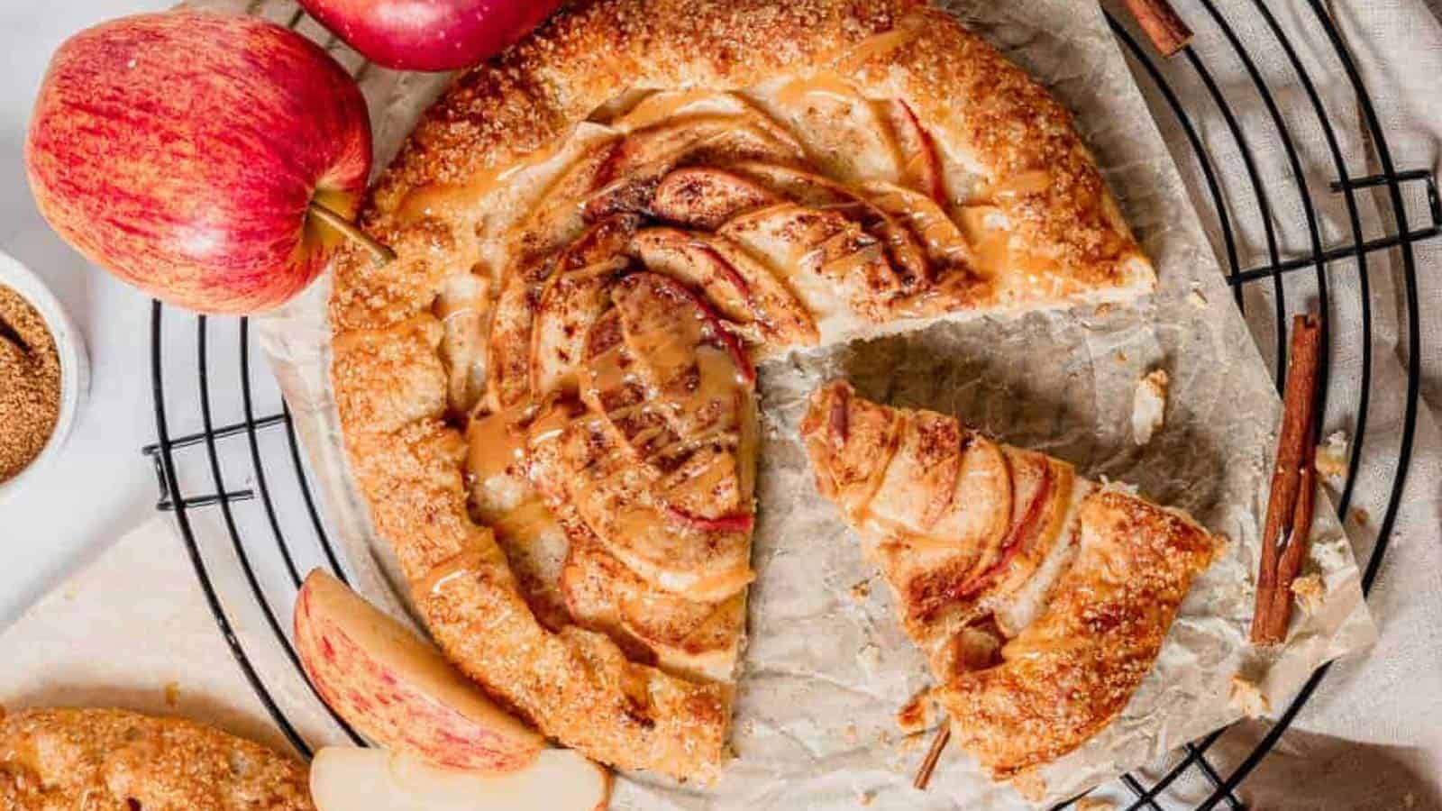A freshly baked apple galette is placed on a wire cooling rack. The galette is golden brown with a slice cut out, revealing tender apple slices arranged in a spiral pattern. Surrounding the galette are whole apples, apple slices, cinnamon sticks, and a bowl of sugar.