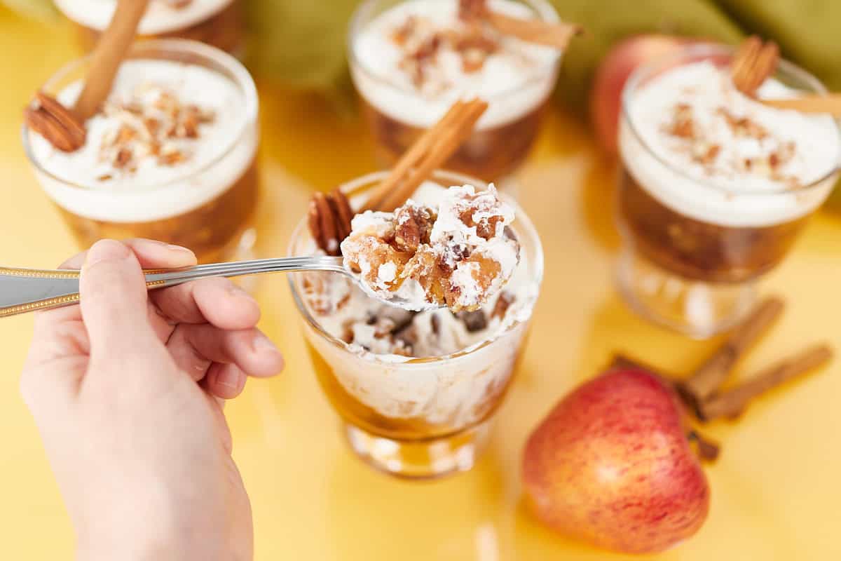 A hand holding a spoonful of parfait topped with whipped cream, nuts, and a cinnamon stick. Additional parfait glasses with similar toppings are in the background, along with a red apple on a bright yellow surface.