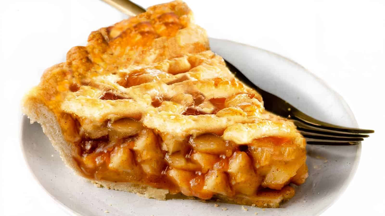 A slice of apple pie, showcasing a golden-brown lattice crust, is served on a white plate with a fork beside it. The pie filling is visible, revealing chunks of apple in a syrupy sauce. The background is white and clean.