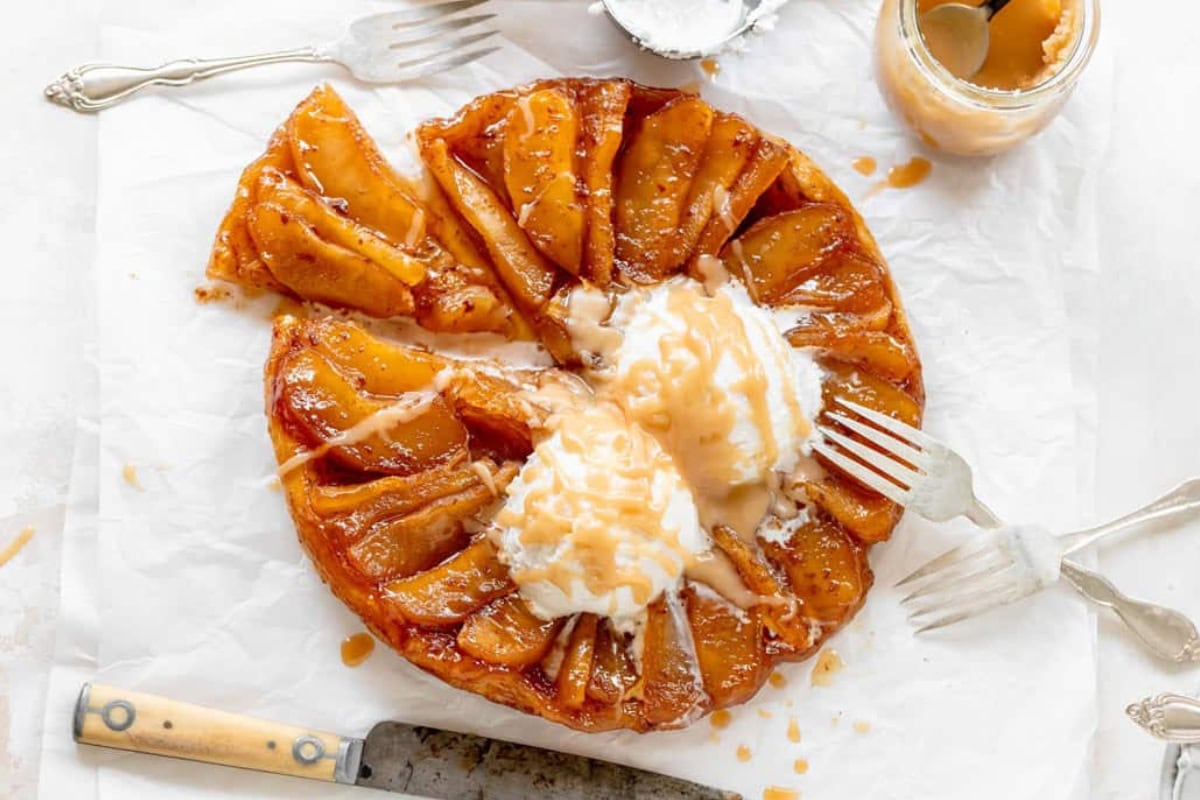 A caramel apple tart topped with two scoops of vanilla ice cream and drizzled with caramel sauce. The tart is on a piece of parchment paper, surrounded by a fork, knife, and a jar of caramel sauce. Two forks are placed into the tart, ready for serving.