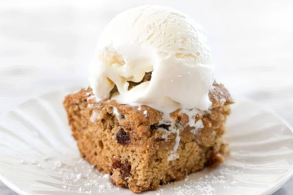 A slice of moist cake with visible pieces of nuts and fruit on a white plate, topped with a generous scoop of vanilla ice cream. The ice cream is slightly melting, forming a creamy cascade over the cake. The background is softly blurred.