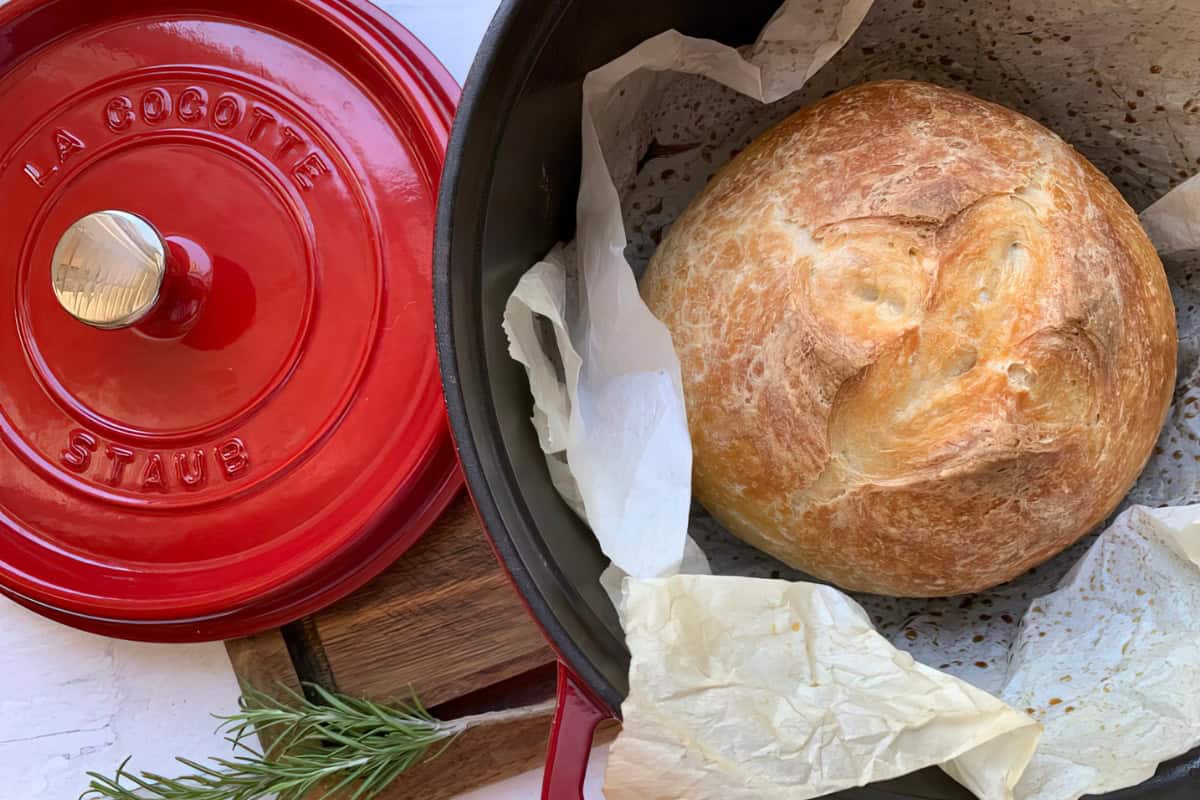 A round loaf of bread with a golden, crusty surface is inside a black dutch oven lined with parchment paper. Next to the dutch oven is a red lid with "La Cocotte Staub" written on it. A sprig of rosemary lies beside the dutch oven.