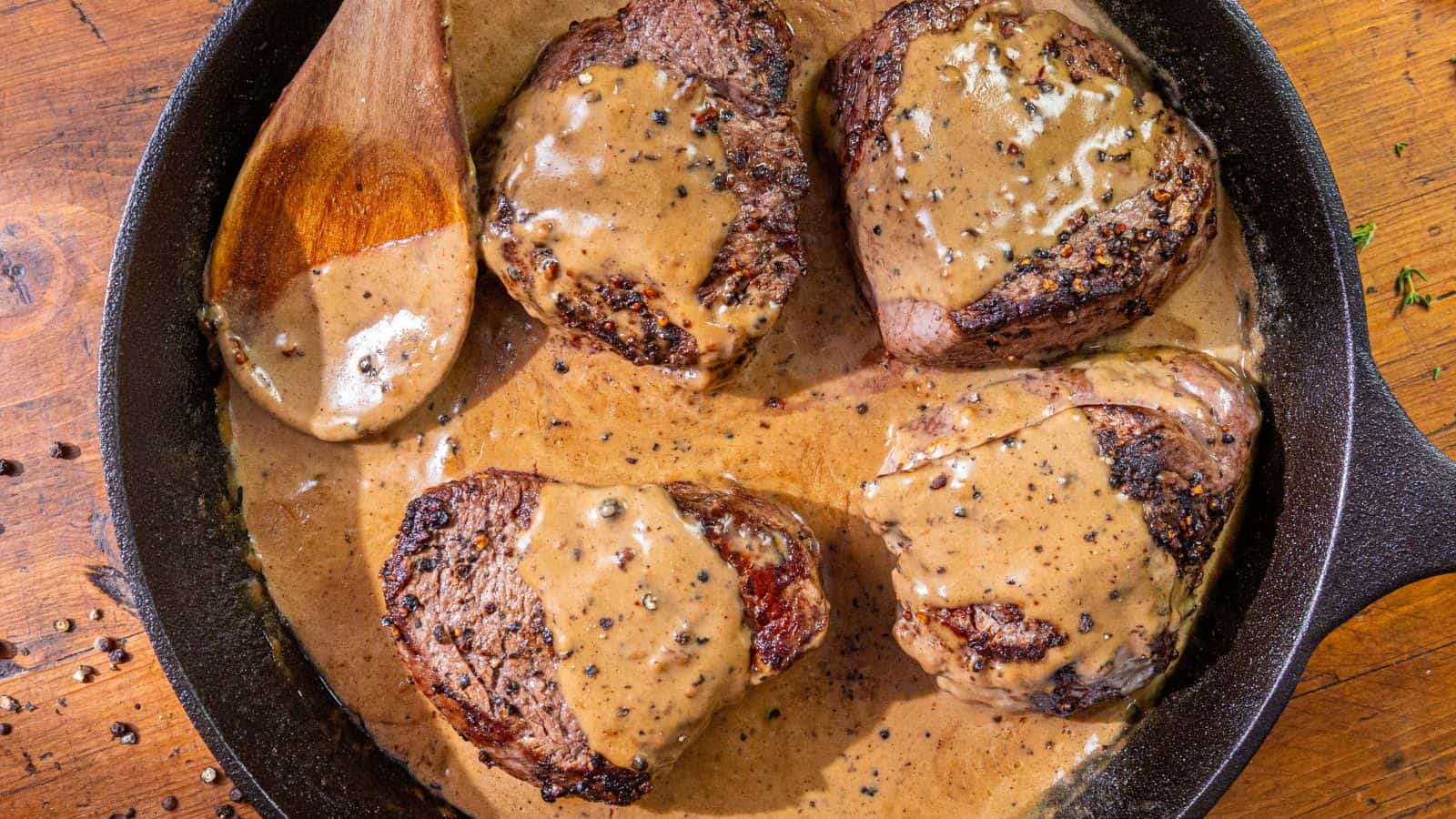 Four cooked steak filets in a cast iron skillet, covered in creamy pepper sauce, with a wooden spoon resting in the sauce.