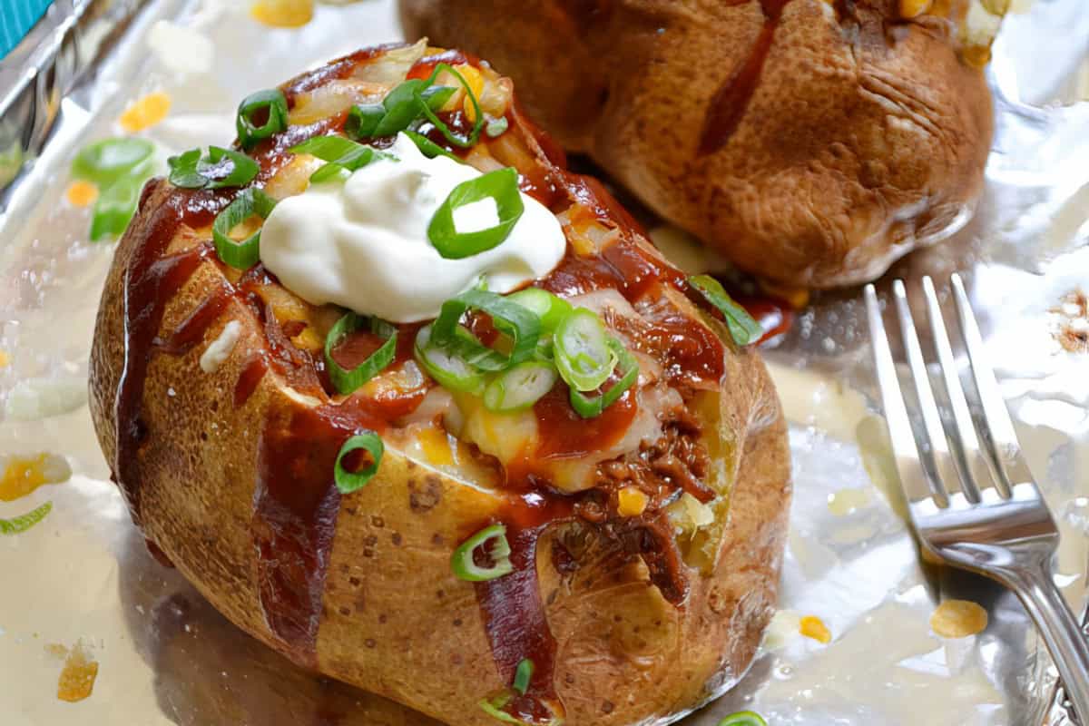 A baked potato topped with sour cream, scallions, shredded cheese, and a drizzle of barbecue sauce sits on a foil-lined surface. A fork is placed nearby. Another baked potato is partially visible in the background.