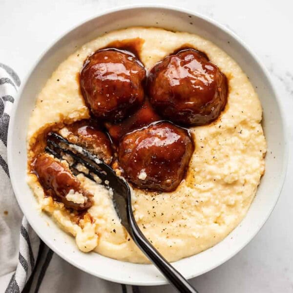A bowl of creamy grits topped with four glazed meatballs and brown sauce, with a fork resting inside the bowl.