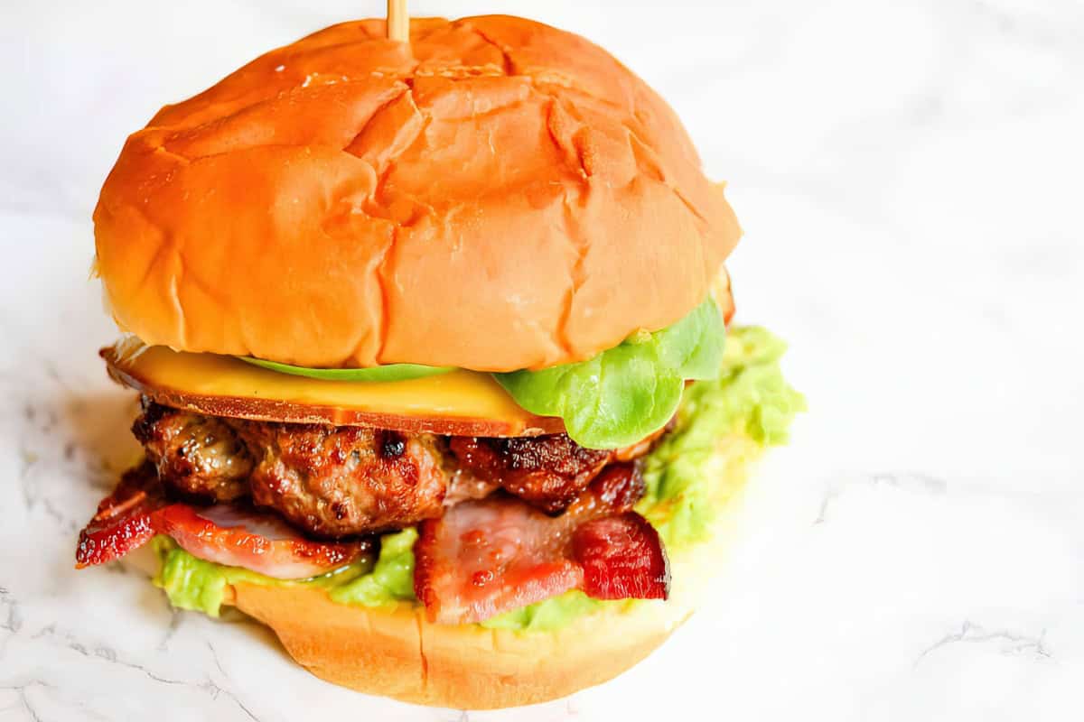 A close-up of a juicy burger with a thick beef patty, crispy bacon, melted cheese, fresh lettuce, and a soft, golden-brown bun. The burger is placed against a white background.