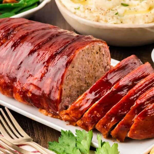 Sliced glazed meatloaf on a white platter with bowls of mashed potatoes, green beans, and a dish of sauce nearby.