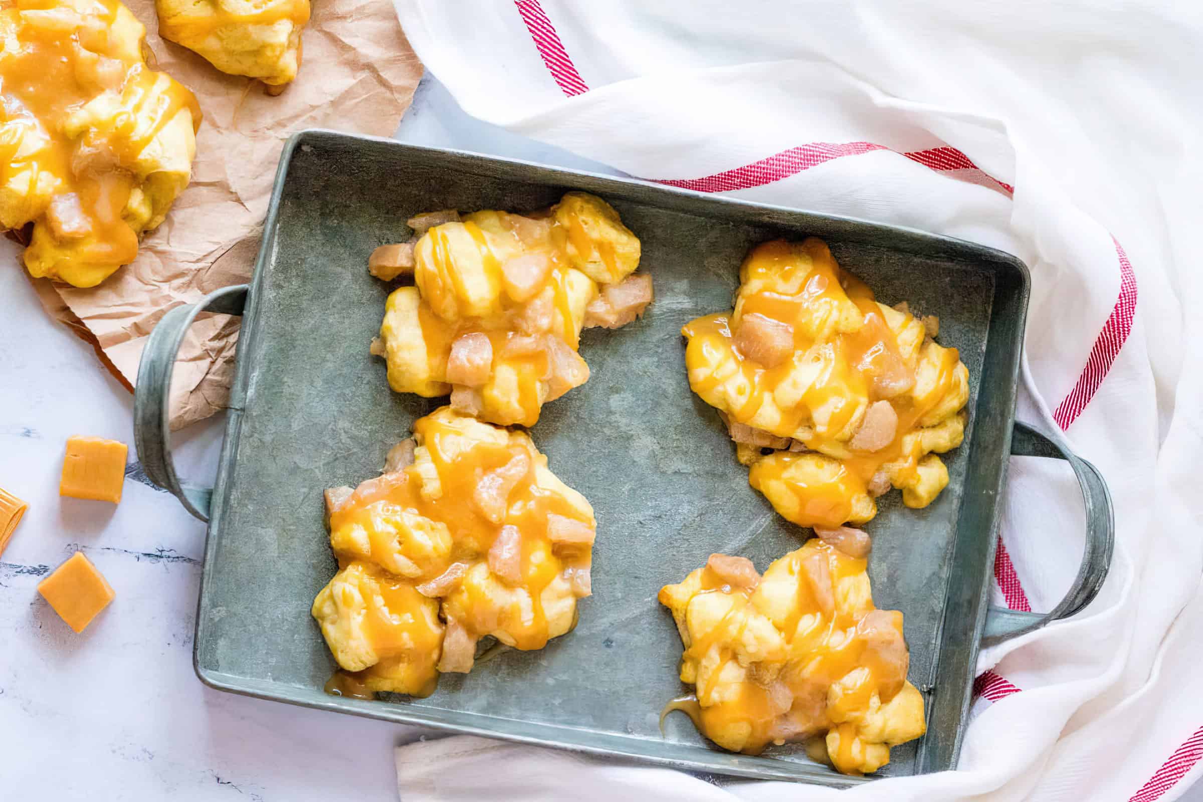 A baking tray with clusters of yellow caramel candy with chewy pieces on a marble countertop. Some candies are on a crumpled piece of parchment paper beside the tray. A white cloth with red stripes is partially visible in the background.