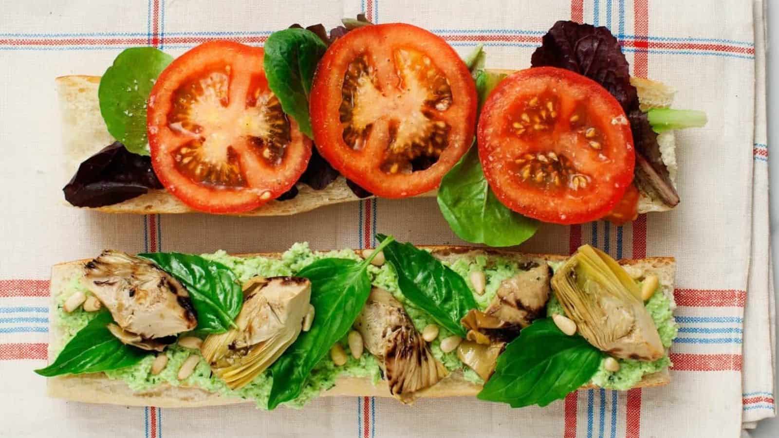 Two sandwich halves on a checkered cloth: one with lettuce and tomato, the other with artichoke, basil, pine nuts, and green spread.