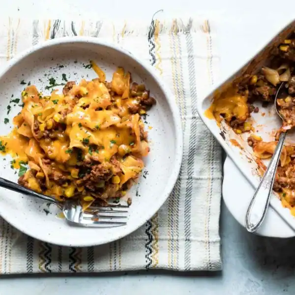 A bowl of pasta with ground meat, corn, and cheese is beside a casserole dish with a serving spoon on a striped cloth.