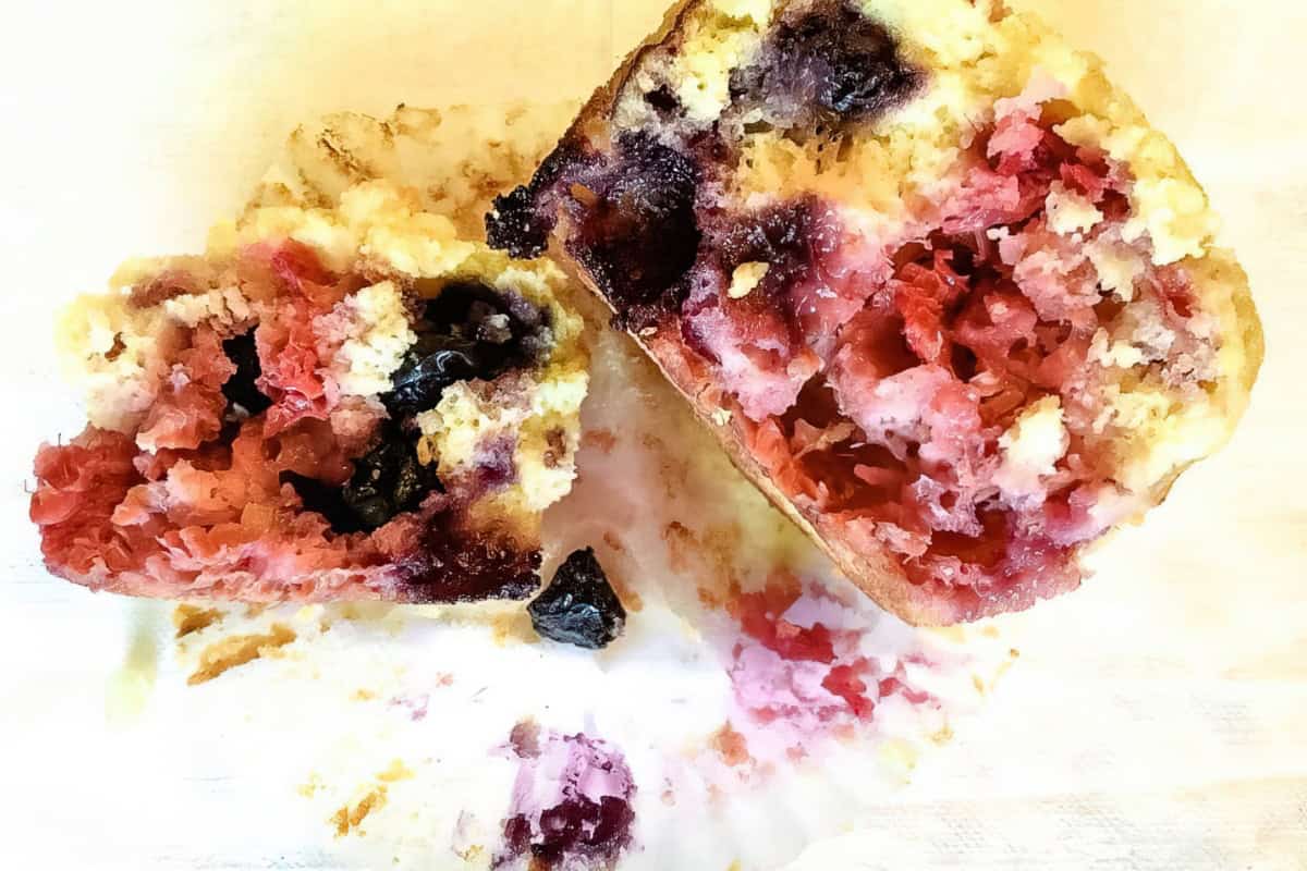 Close-up of a sliced muffin revealing a moist and crumbly interior with mixed berries, including blueberries, raspberries, and possibly blackberries. The muffin sits on a white surface with crumbs and bits of the berries scattered around.