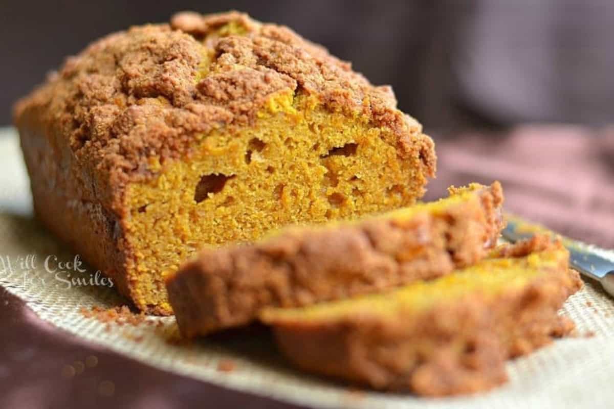A close-up of a loaf of pumpkin bread topped with a crumbly streusel. The loaf is partially sliced, revealing a moist and dense interior. The bread sits on a rustic burlap cloth against a brown background.