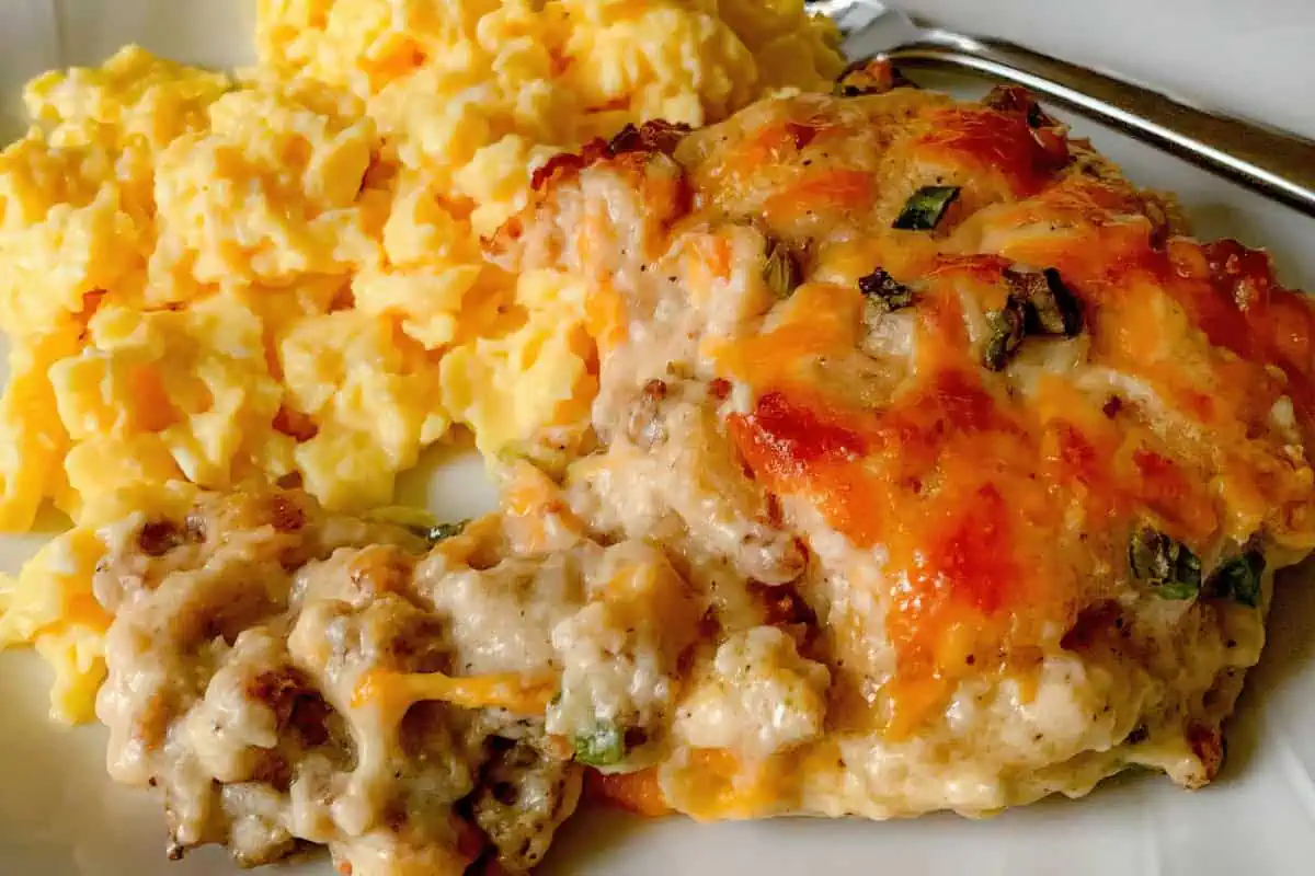 A plate of cheesy sausage and biscuit casserole. The casserole is topped with melted cheese and garnished with green onions, accompanied by scrambled eggs, and served on a white plate with a fork.