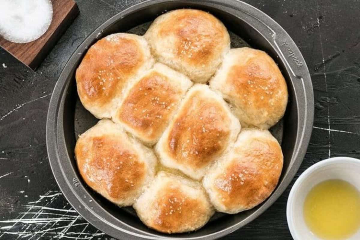 A round baking dish filled with seven freshly baked, golden brown dinner rolls, sprinkled with salt. The dish is set on a dark surface with a small bowl of melted butter and a container of salt nearby. Dough remnants and flour are scattered on the surface.