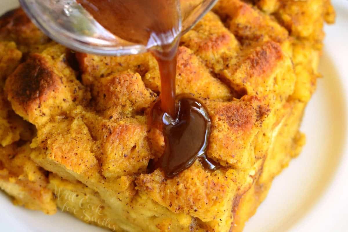 A close-up shot of a golden-brown bread pudding with cinnamon being drizzled with caramel sauce from a glass container. The bread pudding has a crispy, textured surface and is served on a white plate.