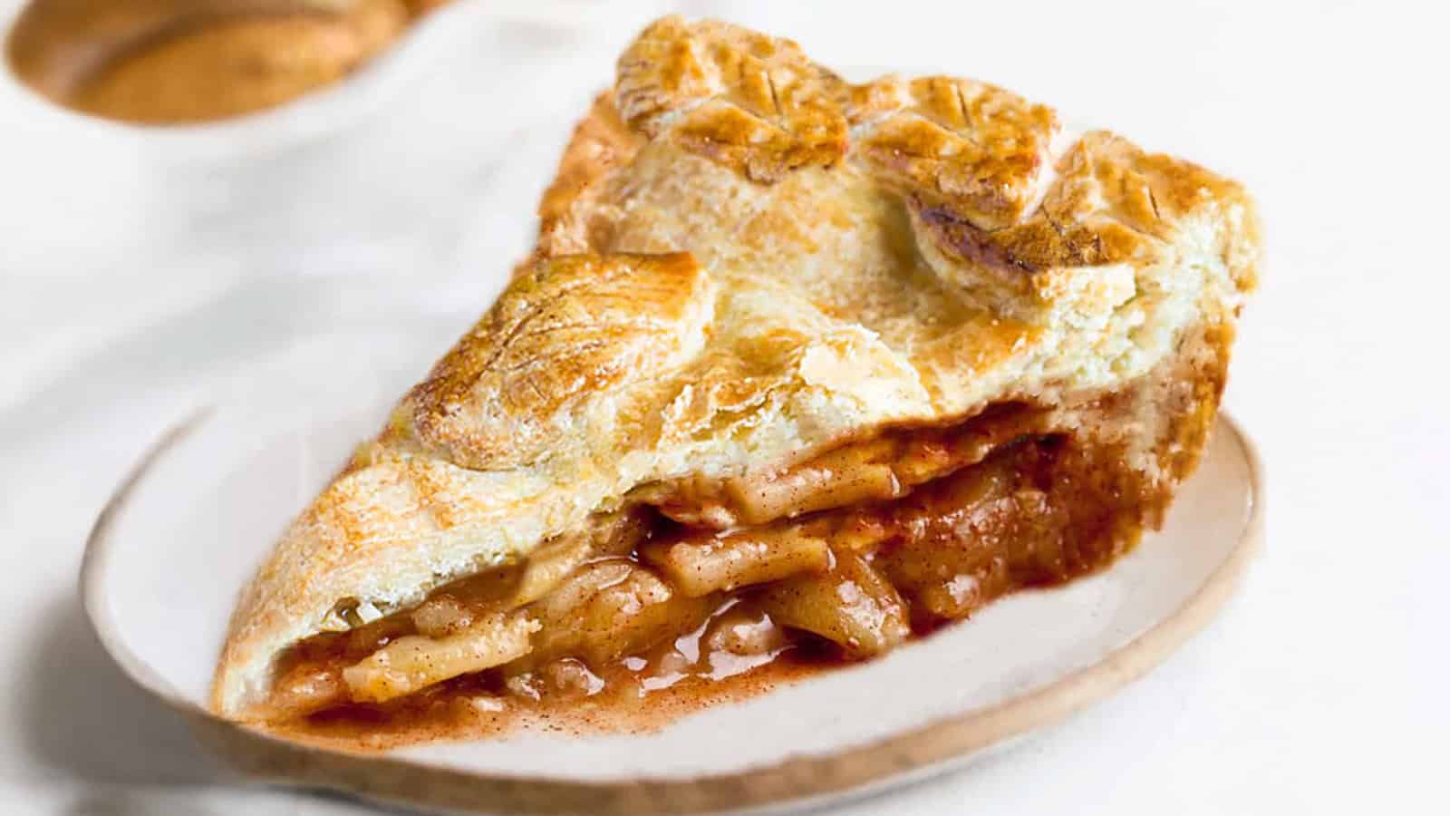 A slice of apple pie with a golden-brown, flaky crust sits on a round white plate. The filling is visible, consisting of sliced apples in a thick, cinnamon-spiced sauce. The pie crust features decorative leaf-shaped cutouts. The background is white.