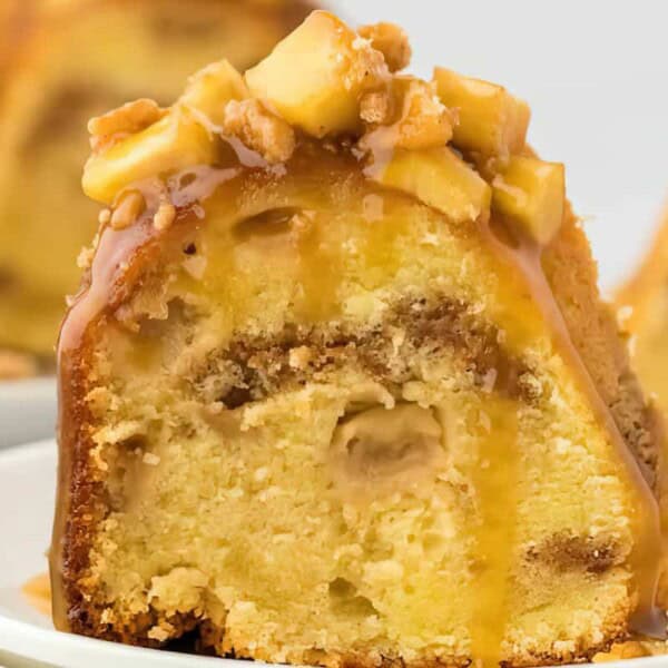 A slice of bundt cake topped with caramel sauce, chopped apples, and walnuts on a white plate.