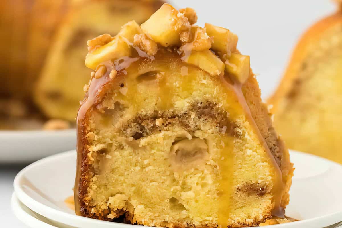 A slice of caramel apple bundt cake on a white plate. The cake is drizzled with caramel sauce and topped with pieces of cooked apple and walnuts. The interior of the cake is moist and appears to have swirls of apple filling.