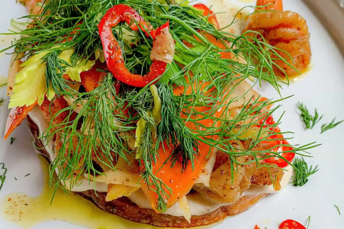 A colorful open-faced sandwich with shrimp, red bell peppers, fresh herbs, and leafy greens on toast. Drizzled with a light dressing, it has a vibrant and fresh appearance, highlighting the textures and colors of the vegetables and seafood.