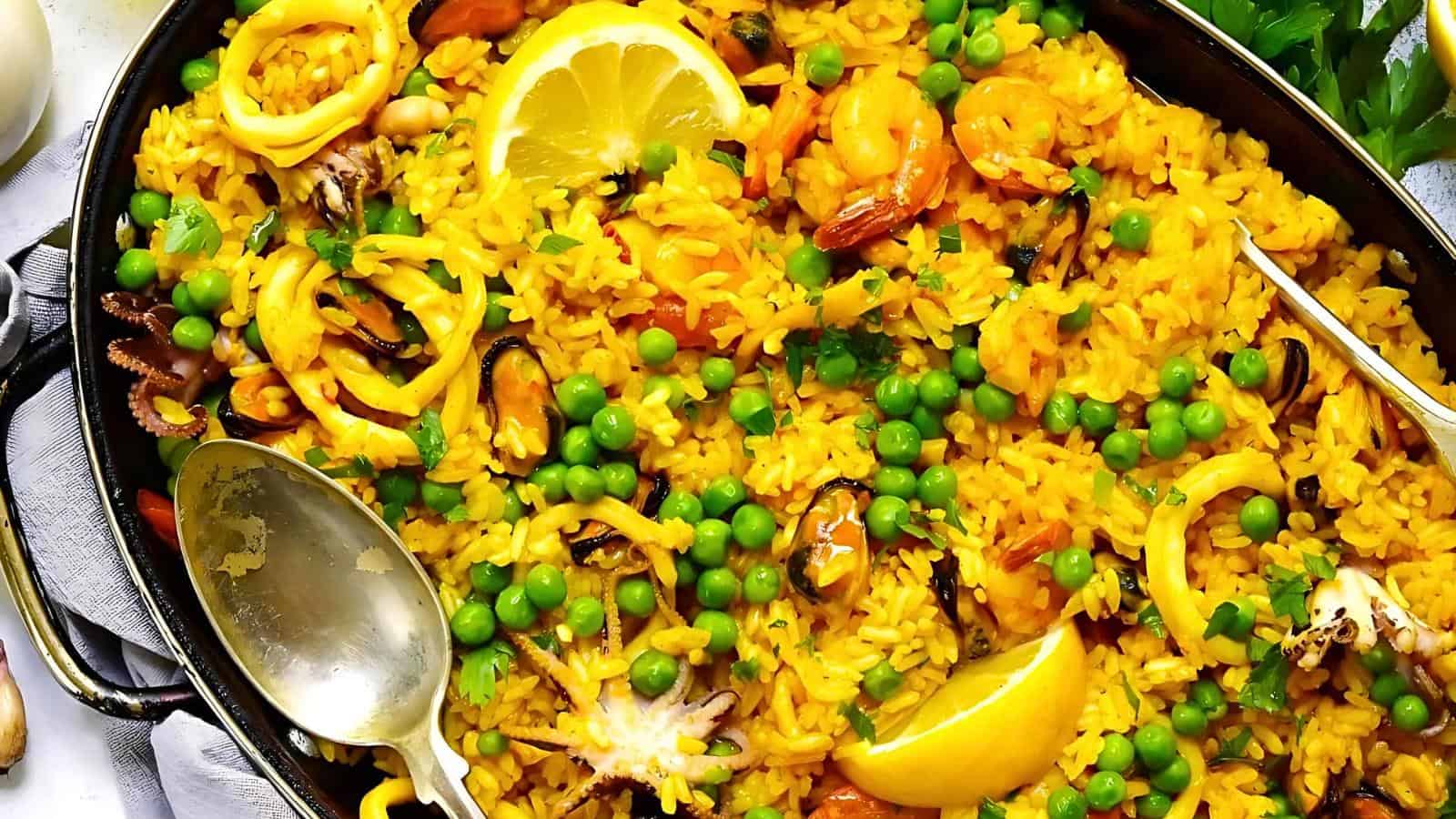 A close-up of a dish of yellow rice, garnished with green peas, shrimp, mussels, calamari, and lemon wedges, served in a black pan. Two utensils, a spoon and a fork, are placed on top of the rice. Fresh parsley is visible in the background.