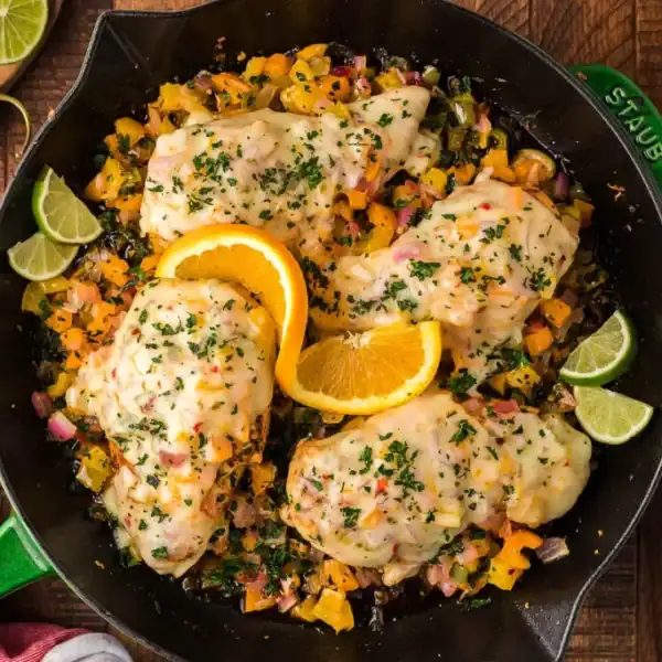 Four baked chicken breasts in a skillet with chopped bell peppers, onions, melted cheese, orange slices, lime wedges, and herbs.